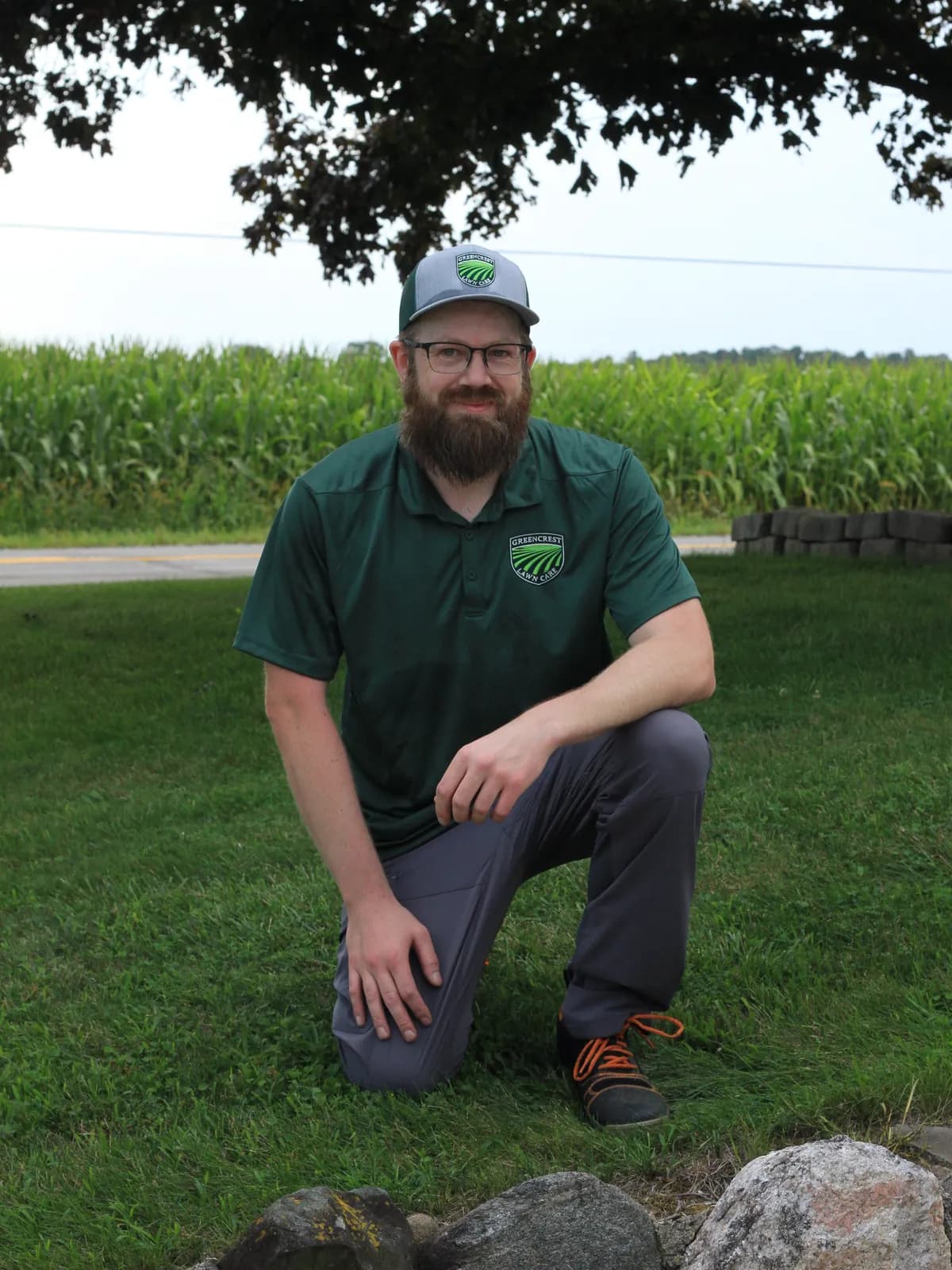 Chris, the owner of Greencrest Lawncare, kneeling on a mown lawn in front of a corn field
