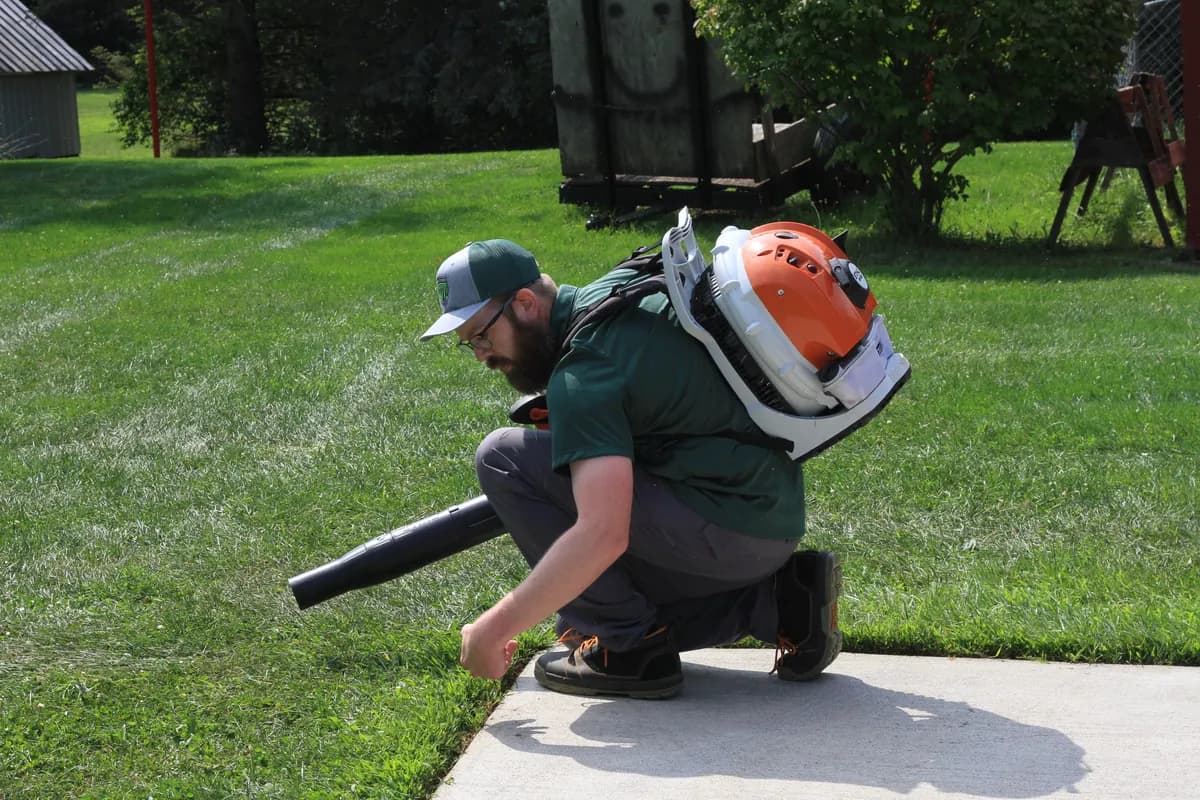Chris kneeling with a backpack blower running, clearing the edge where the lawn meets a walk