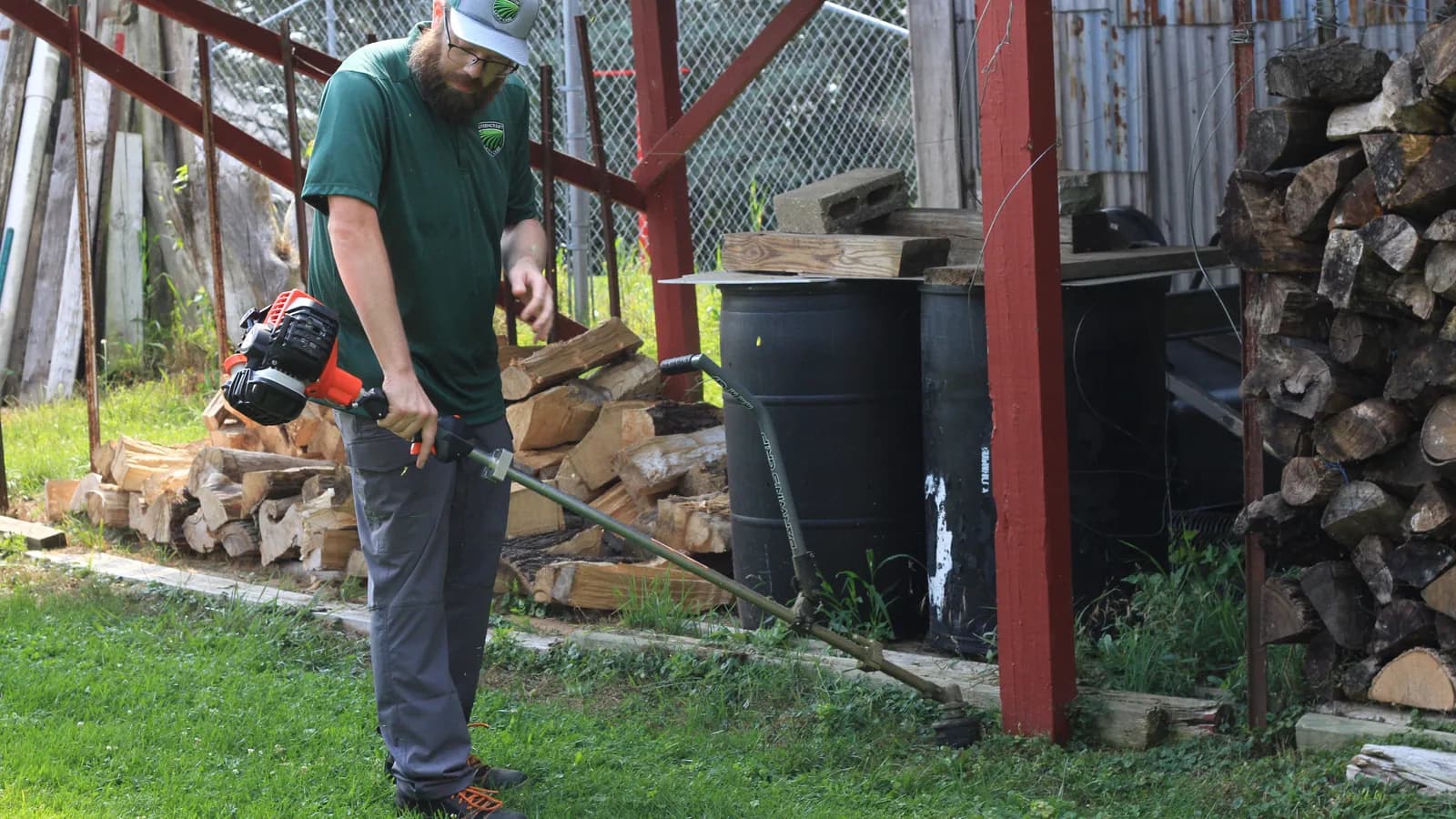 Chris clearing overgrowth with a string trimmer along a woodpile and outbuilding